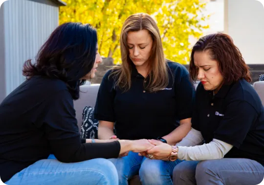 Three women sitting together outdoors, holding hands and praying with eyes closed.