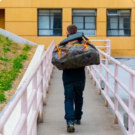 Person walking up a ramp carrying a large camouflage duffel bag and pillow toward a yellow building.