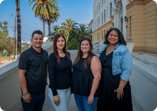 Four people standing outside a large building with palm trees in the background, smiling at the camera.