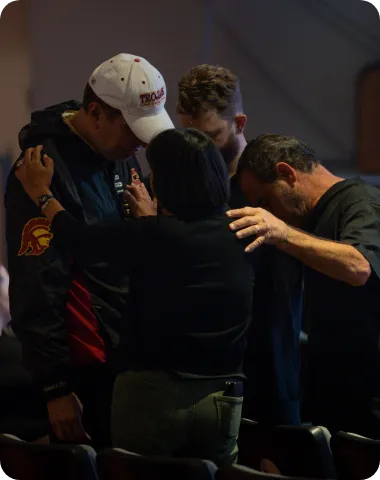 Group of people standing in prayer with heads bowed and hands on each other’s shoulders.