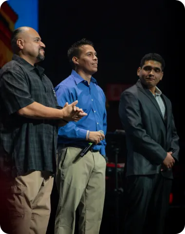 Three men standing on stage dressed in formal attire, one holding a microphone.