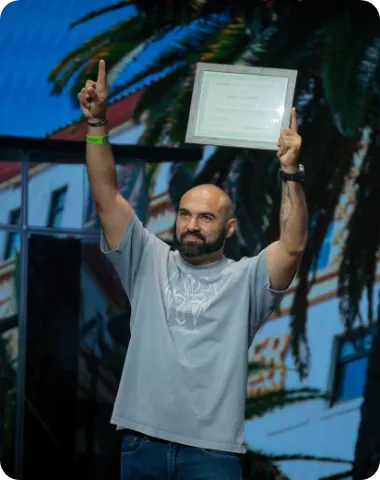 Man holding up a framed certificate on stage, smiling and pointing upward.