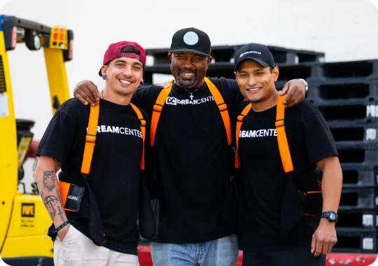 Three men wearing black Dream Center shirts and orange safety straps smiling together outdoors.